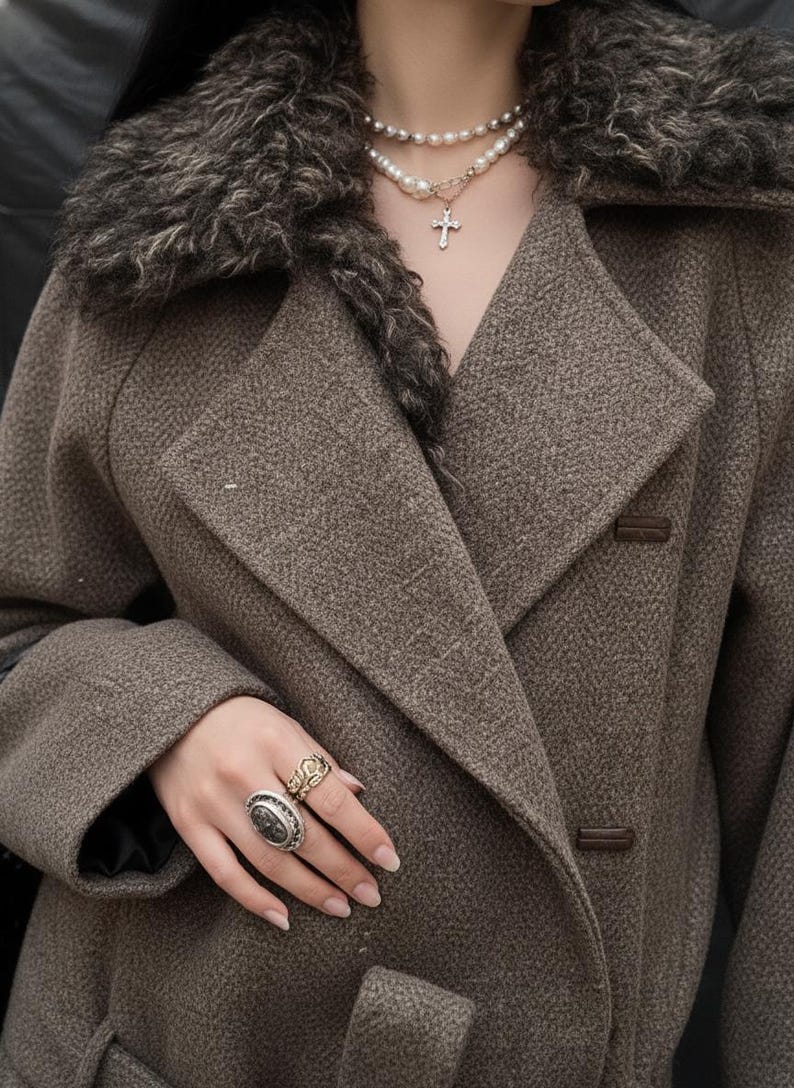 May include: A brown wool coat featuring a faux fur collar. The coat has a double-breasted design with large lapels and two brown buttons. A pearl necklace with a cross pendant and multiple rings are also visible.