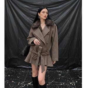 May include: A brown wool coat featuring a faux fur collar and a belted waist. The coat has a peplum hem and is worn with black knee-high boots. A black handbag is worn over the shoulder. The background is a black backdrop.