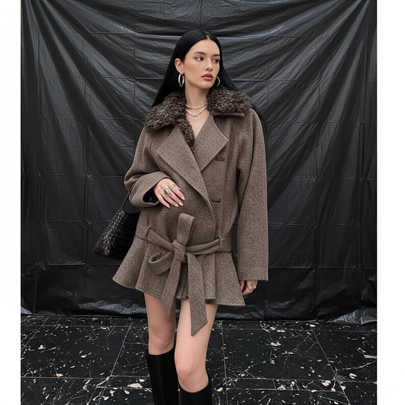 May include: A brown wool coat featuring a faux fur collar and a belted waist. The coat has a peplum hem and is worn with black knee-high boots. A black handbag is worn over the shoulder. The background is a black backdrop.
