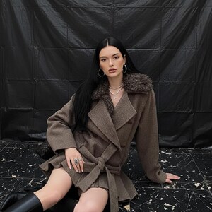 May include: A brown wool coat with a faux fur collar and a belted waist. The coat is knee-length and has a double-breasted design. The model is wearing black boots and silver hoop earrings.