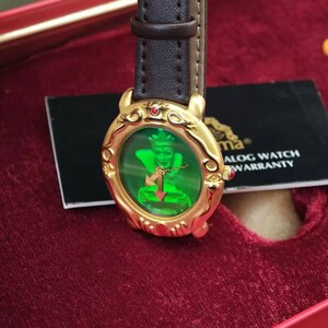 May include: A gold wristwatch with a green face showing a figure and a red arrow. It has a brown leather strap and a small red gem. The watch is presented in a red box.
