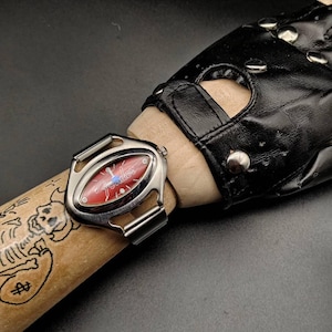 May include: A silver-toned Fender watch with a red face and the Fender logo. The watch is on a wooden dowel with a skull and money bag illustration. A black leather fingerless glove with silver studs is also visible.