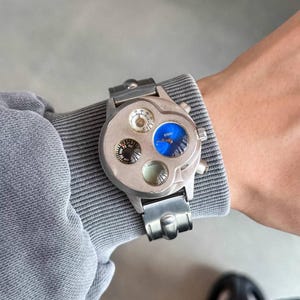 Puede incluir: Un reloj de pulsera plateado con múltiples esferas, incluyendo una brújula y un indicador azul. El reloj se lleva sobre una camisa gris de manga larga. El reloj tiene un diseño industrial único.