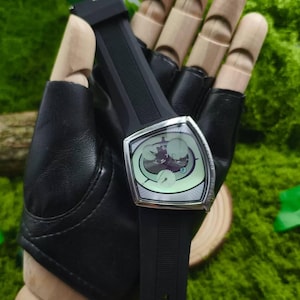 May include: A black wristwatch with a unique, angular silver case and a black rubber strap. The watch face has a light green background with a detailed graphic. The watch is held by a black leather glove.