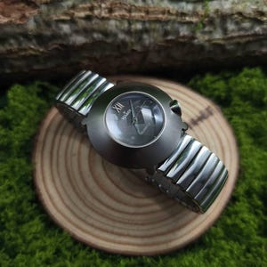 Op de afbeelding: Een zilverkleurig polshorloge met een donkere wijzerplaat, met de tekst "Vidage Quartz". Het horloge heeft een geribbelde metalen band en wordt gepresenteerd op een klein houten schijfje, tegen een achtergrond van groen mos en een boomstam.