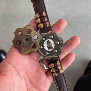 Puede incluir: Un reloj de pulsera único con una correa de cuero marrón y detalles en latón. La esfera del reloj presenta un diseño de diana, y la correa está adornada con detalles en forma de bala. El reloj está abierto, revelando un cilindro giratorio con el texto "357 MAG".
