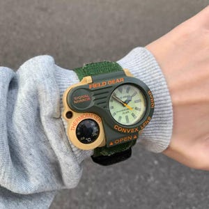 May include: A green and tan Field Gear watch with a thermometer and signal mirror. The watch has a green fabric strap and a round face with the text "ALBA" and "CONVEX LENS". The watch also has a thermometer that reads 20 degrees Celsius.