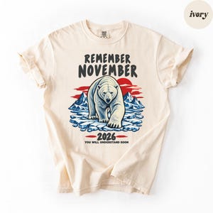 May include: An ivory t-shirt featuring a graphic of a polar bear walking on ice floes with mountains and a red sun in the background. The text reads REMEMBER NOVEMBER 2026 YOU WILL UNDERSTAND SOON in dark gray lettering above and below the illustration.
