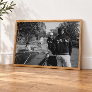 May include: A black and white framed photograph depicting a person wearing a dark jacket with the word TIGERS printed on the back, standing near a convertible car while a police officer stands nearby on a tree-lined street.
