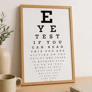 May include: A framed eye test chart with a wooden frame. The chart features the text "EYE TEST" at the top, followed by a humorous message. A mug and vase are visible in the background. The text reads: "IF YOU CAN READ THIS YOU ARE SITTING ON THE TOILET..."