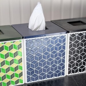 May include: A row of four decorative tissue box covers. Each box has a different geometric pattern in green, blue, black, and pink. The boxes are square with a dark top and a square opening for tissue dispensing.