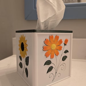 May include: White tissue box with a black top and floral designs. The box features a sunflower and an orange flower with green leaves. White tissues are pulled from the top of the box.