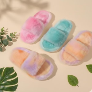 Puede incluir: Cuatro zapatillas esponjosas de punta abierta en tonos rosa, turquesa y tie-dye naranja-morado. Las zapatillas tienen una textura suave y afelpada y una correa ancha en la parte superior. El fondo es de color beige claro.