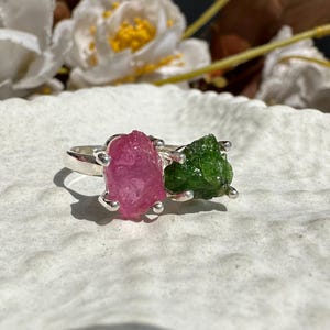 May include: A silver ring featuring two raw gemstones: a vibrant pink stone and a green stone. The stones are set side-by-side, creating a unique and natural design. The ring is displayed on a textured white surface.