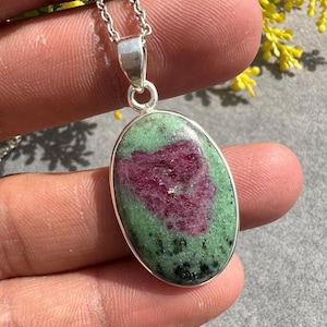 May include: Oval pendant necklace with a natural ruby zoisite stone. The stone has a green base with a heart-shaped ruby centre. The pendant is set in silver-coloured metal and hangs from a delicate chain.