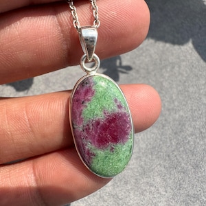 May include: An oval-shaped pendant featuring a polished ruby and zoisite gemstone with vibrant magenta and green mottled patterns. The stone is set in a smooth silver-toned metal bezel and hangs from a matching silver-toned link chain necklace.