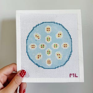 May include: A square, hand-stitched needlepoint canvas featuring a light blue circle with a dark blue outline. The circle is filled with small, colorful, patterned shapes in red, green, and yellow. The initials "ML" are in the bottom right corner.