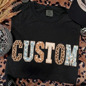 Könnte beinhalten: Schwarzes T-Shirt mit dem Wort "CUSTOM" in einem Patchwork-Design. Die Buchstaben zeigen Leopardenmuster, Blumenmuster und ein Karomuster. Das T-Shirt befindet sich auf einem Leopardenmuster-Hintergrund.