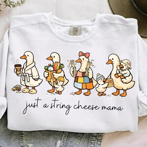 May include: White sweatshirt featuring a whimsical illustration of a family of ducks. The ducks are depicted in various outfits, carrying items like coffee, groceries, and string cheese. The text "just a string cheese mama" is written in cursive below the ducks.