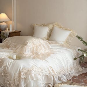 May include: A cream-colored bedding set featuring a duvet cover and two square pillow shams. The fabric is adorned with intricate floral lace trim and layered ruffles along the edges, creating a textured appearance on the bed linens.