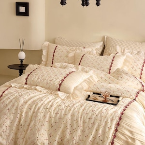 May include: A cream-colored bedding set featuring a quilted texture with a delicate diamond pattern and small red floral accents. The set includes ruffled pillow shams and a matching comforter with decorative trim, creating a layered appearance.