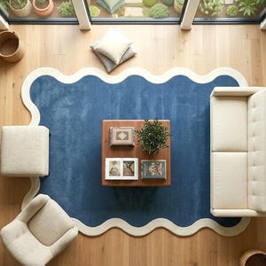 Könnte beinhalten: Draufsicht auf ein Wohnzimmer mit einem blauen Teppich mit weißem Wellenrand. Ein cremefarbenes Sofa, ein Stuhl und ein Hocker umgeben einen Holztisch. Der Tisch zeigt Bücher und eine kleine Pflanze. Natürliches Licht fällt ein.