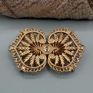 May include: A gold-tone metal cloak clasp featuring two symmetrical, mirrored halves. Each side displays an intricate, openwork leaf and scroll pattern radiating from a central, textured circular floral motif, creating a decorative fastener.