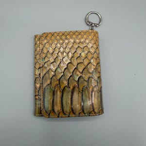 May include: A tri-fold wallet with a key ring, featuring a snakeskin pattern in shades of tan, brown, and green. The wallet has a textured surface and a silver-colored key ring attached to the top.