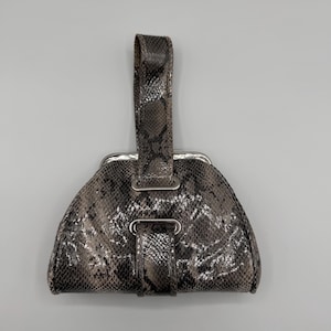 Peut inclure: Un petit sac à main semi-circulaire avec un motif peau de serpent dans les tons de gris et de marron. Il est doté d'une poignée et d'un fermoir argenté. Le sac a une fermeture à lanière avec un anneau argenté.