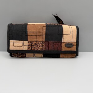 Puede incluir: Una cartera de patchwork con una variedad de texturas y patrones. La cartera presenta cuadrados de tela negra, marrón claro y estampado de leopardo. Un borde negro rodea la cartera y una pequeña etiqueta está adjunta.