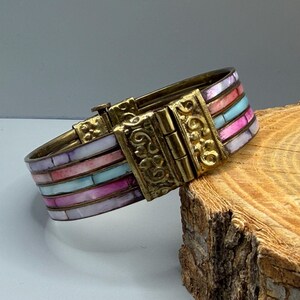 Peut inclure: Un bracelet articulé avec une finition en laiton et des incrustations rectangulaires colorées. Les incrustations présentent des nuances de rose, de bleu et de blanc. Le fermoir a un design orné. Le bracelet repose sur un morceau de bois.
