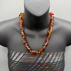 Puede incluir: Un collar con grandes cuentas rectangulares de color ámbar con espaciadores plateados. Las cuentas tienen un aspecto veteado. El collar se exhibe en un maniquí negro.