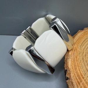 May include: A stretch bracelet featuring alternating smooth, white, rectangular resin links and polished silver-tone metal bars. The links are rounded at the edges and connected by a flexible internal band, creating a continuous, chunky, geometric design.