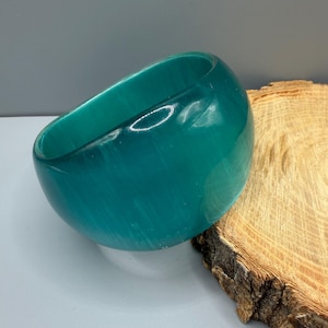 Puede incluir: Un brazalete ancho y translúcido de color verde azulado con un acabado liso y pulido, que presenta sutiles estrías verticales en todo el material. El accesorio circular tiene un perfil exterior redondeado y un centro hueco de tono azul verdoso.
