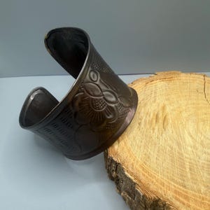 May include: A dark bronze cuff bracelet with a floral design and textured patterns. The wide bracelet has an open design and is displayed on a wooden surface. The cuff is a statement piece of jewellery.