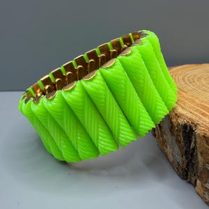 Könnte beinhalten: Ein leuchtend neongrünes Stretch-Armband mit einer Reihe zylindrischer, diagonal gerippter Segmente, die durch einen goldfarbenen Metallrahmen verbunden sind. Die leuchtend grünen Teile sind in einem sich wiederholenden Muster angeordnet.
