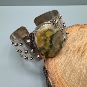 May include: A wide cuff bracelet featuring a large, oval-shaped stone with mottled yellow, grey, and black patterns. The band is made of textured silver-toned metal adorned with numerous small, raised spherical studs along the edges.
