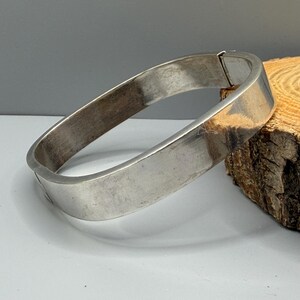 May include: A polished silver-toned metal bangle bracelet with a smooth, wide, and flat surface. The bracelet features a hinged opening mechanism with a visible clasp closure, forming a rectangular shape with rounded corners and a reflective finish.