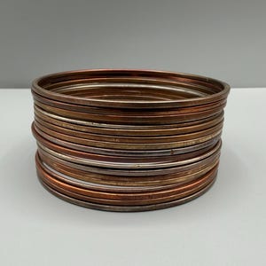 Puede incluir: Una pila de brazaletes metálicos circulares y delgados que presentan bandas alternas en tonos cobre, bronce y plata. Las pulseras están dispuestas en una columna vertical, mostrando un acabado liso y pulido con un diámetro uniforme y constante.