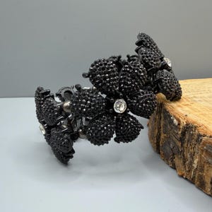 May include: A black cuff bracelet featuring a series of connected flower-shaped links. Each petal is covered in small, dark, textured beads, with a single clear, round rhinestone set in the center of each flower. The bracelet has a dark metal finish.