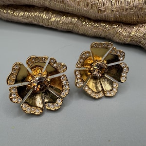 Może przedstawiać: Para złotych kolczyków w kształcie kwiatów. Każdy kolczyk ma centralny brązowy kamień szlachetny, otoczony złotymi płatkami i przezroczystymi kryształami. Leżą na jasnoszarej powierzchni.