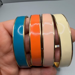 Puede incluir: Cuatro pulseras tipo brazalete con un acabado liso y brillante y bordes de metal dorado. Las pulseras son de color azul cerceta, naranja brillante, rosa pastel suave y crema. Cada brazalete presenta una banda de color sólido en un marco dorado.