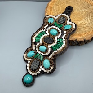 May include: An elongated, dark brown leather cuff bracelet featuring intricate beadwork. It is embellished with smooth, oval-shaped turquoise and malachite stones, interspersed with small white square beads and rows of metallic bronze seed beads.