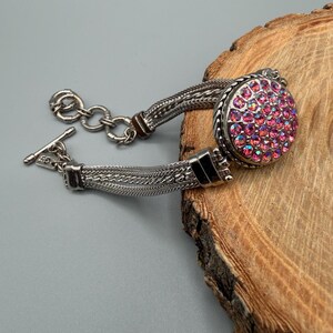 May include: A silver-toned bracelet featuring a multi-strand chain design and a circular focal piece encrusted with numerous small, iridescent pink crystals. The bracelet includes a toggle clasp closure and a small metal tag marked with the letters GS.