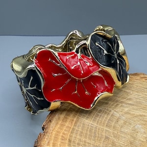 May include: A gold-coloured metal hinged bracelet with red and black enamel leaf designs. The bracelet has a gold trim and is set on a wooden surface.