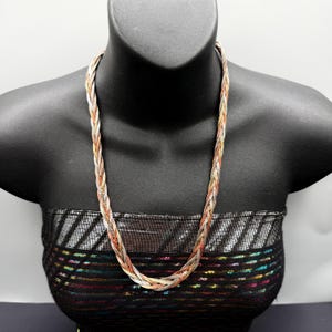 May include: A multi-tone braided necklace in silver, gold, and rose gold. The necklace is displayed on a black mannequin. The chain is made of interwoven strands, creating a textured appearance. A statement piece.