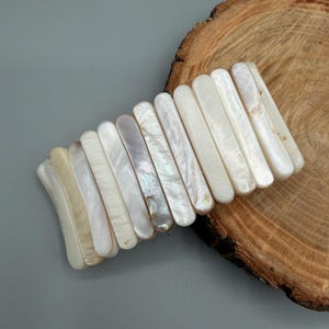 Peut inclure: Un bracelet extensible composé de quatorze segments rectangulaires en nacre polie. Chaque segment présente une surface lisse et irisée avec des nuances variées de blanc crème et des tons nacrés doux disposés en un motif linéaire.