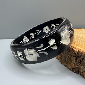May include: A wide, dark black bangle bracelet featuring a smooth, polished surface. The exterior is decorated with inlaid white floral motifs, including stylized roses and delicate leafy vines that wrap around the circumference of the circular band.