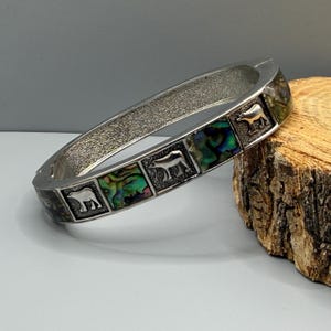 Peut inclure: Un bracelet jonc articulé de couleur argentée composé de panneaux carrés alternés. Le motif présente des silhouettes d'ours en relief et des incrustations de nacre d'ormeau aux reflets verts, bleus et violets sur un métal texturé.