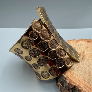 May include: A wide, polished gold-tone metal cuff bracelet featuring a repeating pattern of recessed, circular indentations. Each circular depression contains a textured, granular surface that contrasts with the smooth, reflective gold finish.